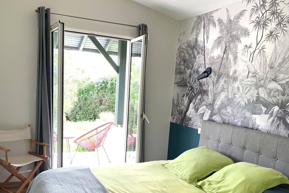 A bedroom with a bed, a chair, a view through a glass door, and a wall with a tropical mural.