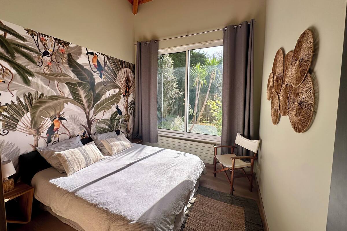 A bedroom with a large bed, a window with curtains, a chair, and a wall with a tropical mural.