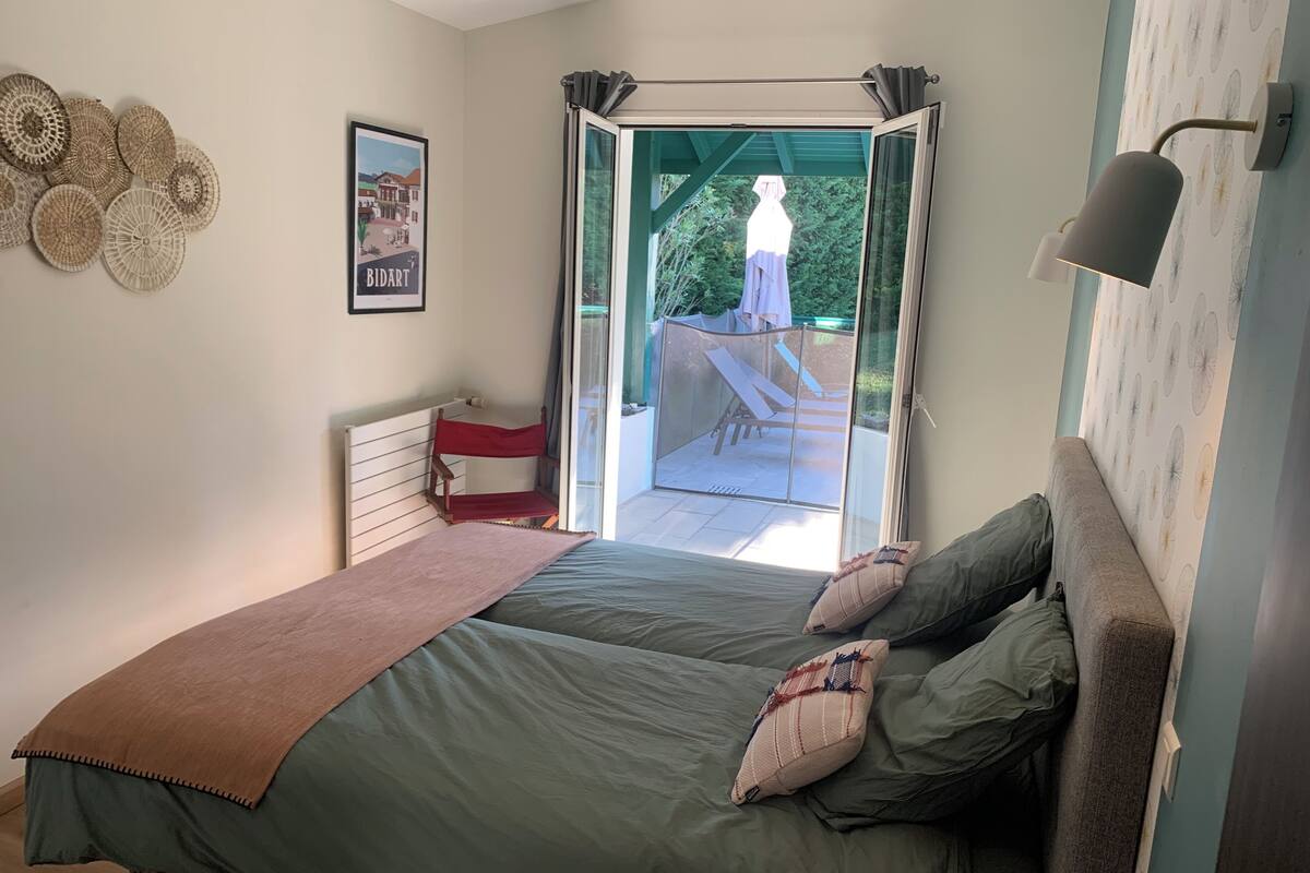 A bedroom with a bed, a bench, a chair, a window, and a wall-mounted lamp.