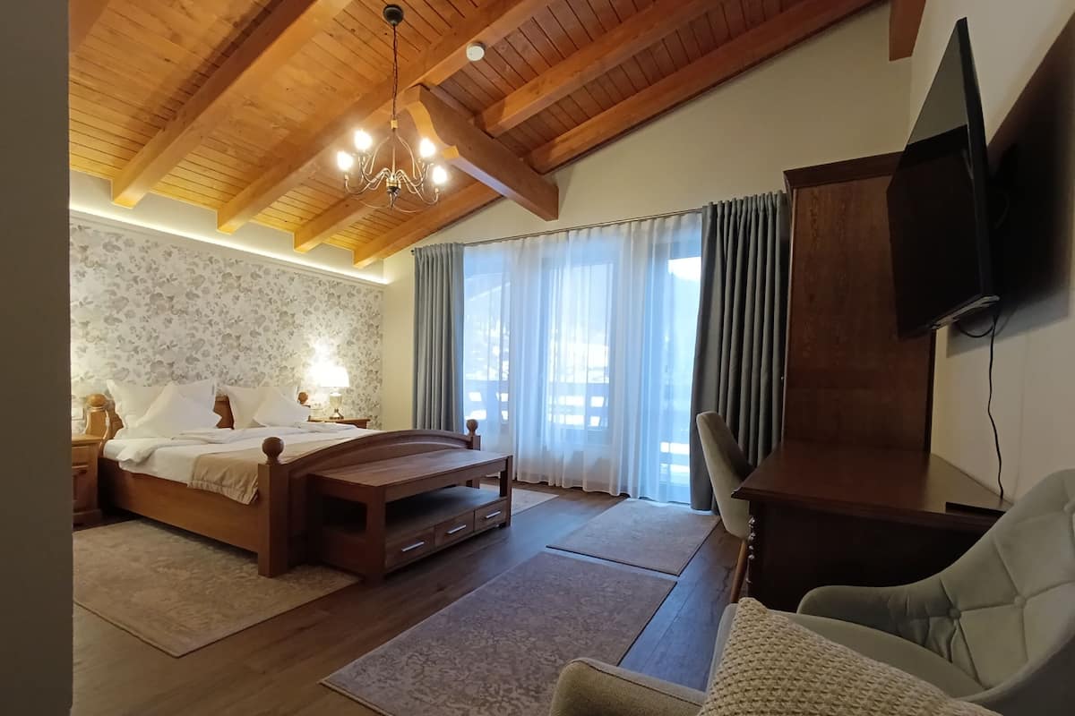 A bedroom with a wooden bed, a desk, a chair, a television, and a large window with curtains.