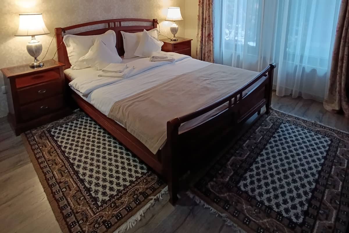 A bedroom with a wooden bed, two bedside tables with lamps, a patterned rug, and a large window with curtains.