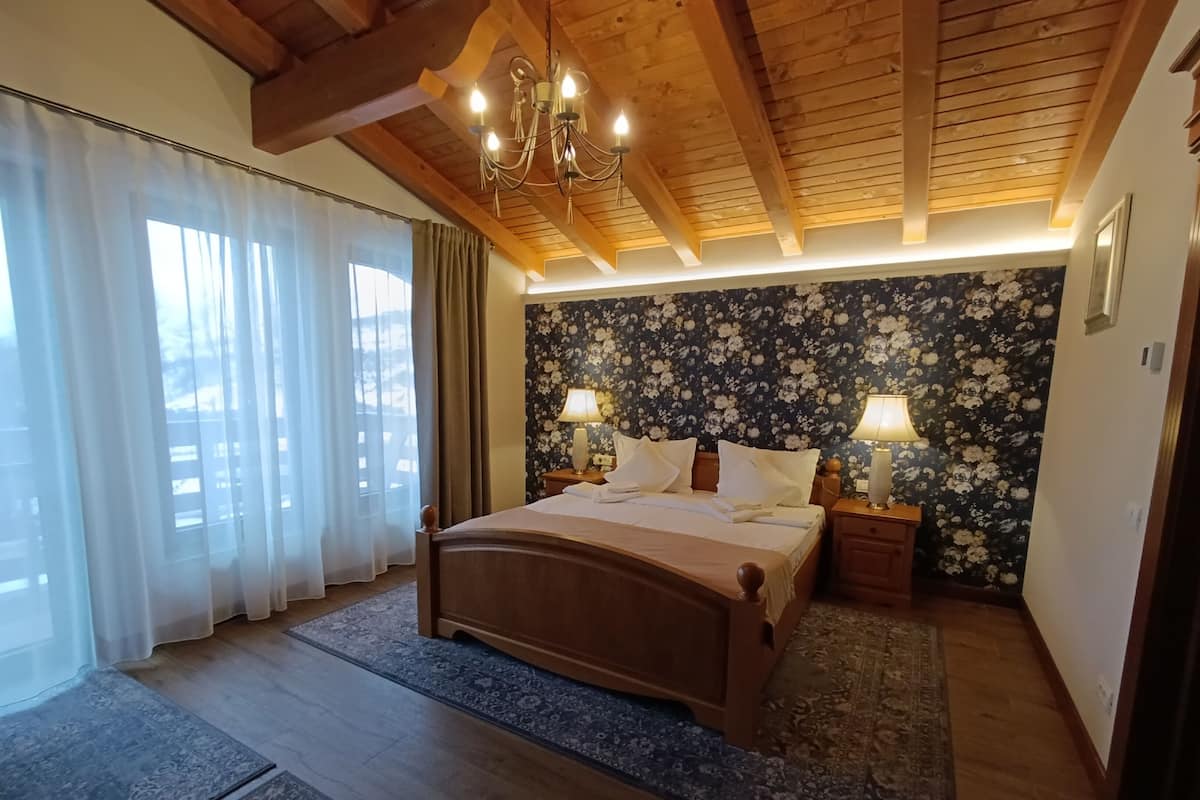 A bedroom with a wooden ceiling, a large bed, two bedside tables with lamps, a floral wallpaper, and a view through sheer curtains.