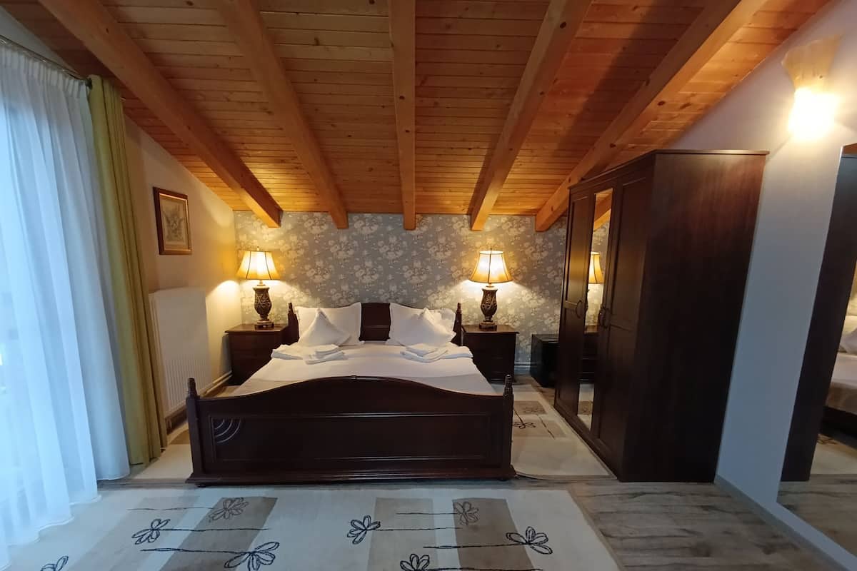 A bedroom with a wooden ceiling, a bed with white linens, two bedside lamps, a floral-patterned wall, and a large wardrobe.