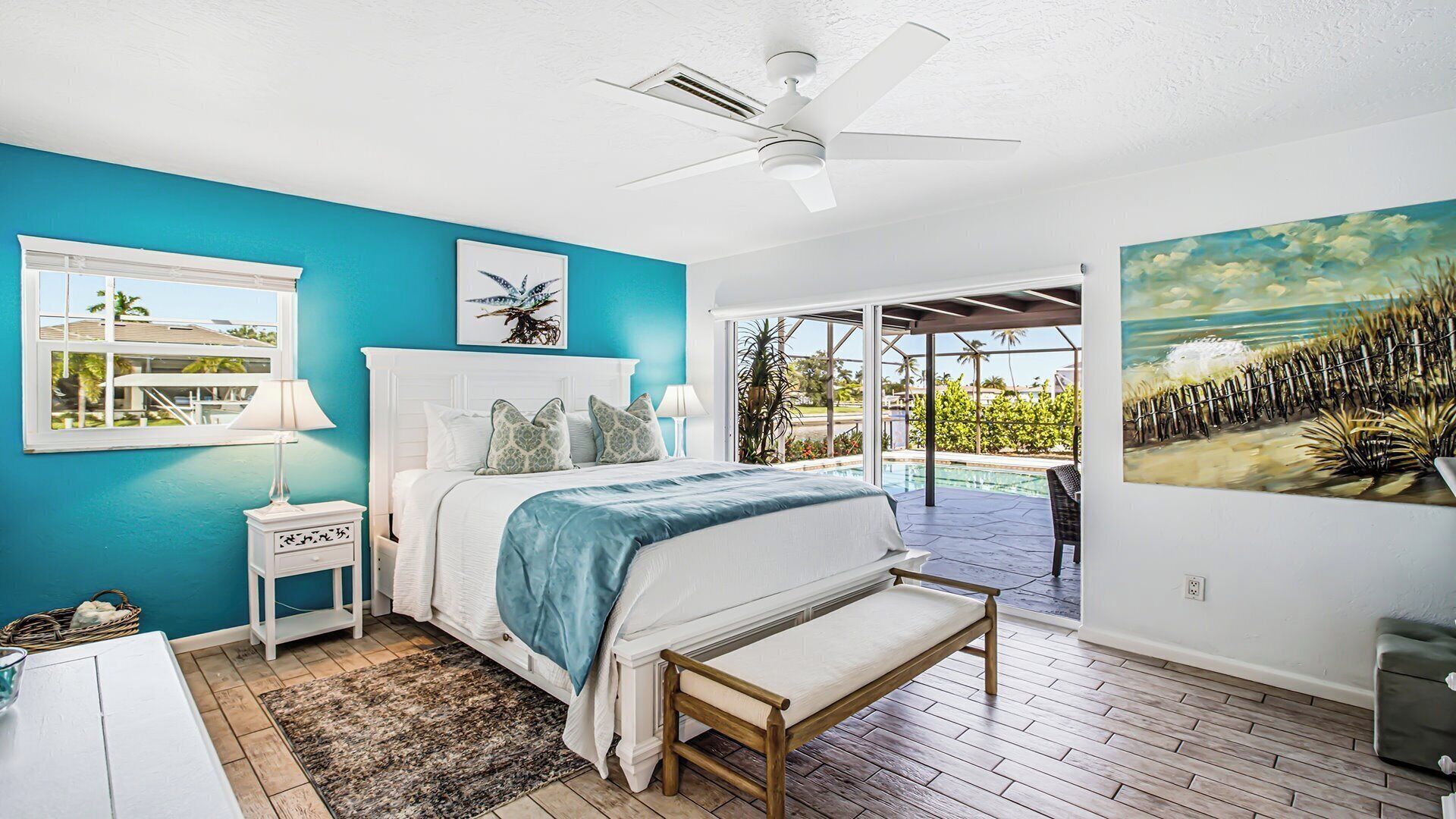 A bedroom with a bed, a ceiling fan, a window, a nightstand, a bedside table, a bench, and a painting.