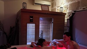 Un bain à remous où se trouvent deux personnes, un meuble en bois doté d’un thermostat et, au fond, un mur en pierre.