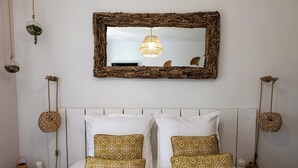Une chambre avec un lit blanc, deux oreillers blancs et deux oreillers à motifs. Un miroir tressé est accroché au-dessus du lit, et une suspension lumineuse est fixée au plafond.