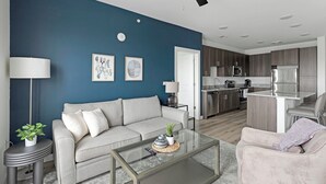 A modern living room with a grey sofa, a glass coffee table, and a kitchen in the background. - Landing Apartments Garland, TX (Garland)