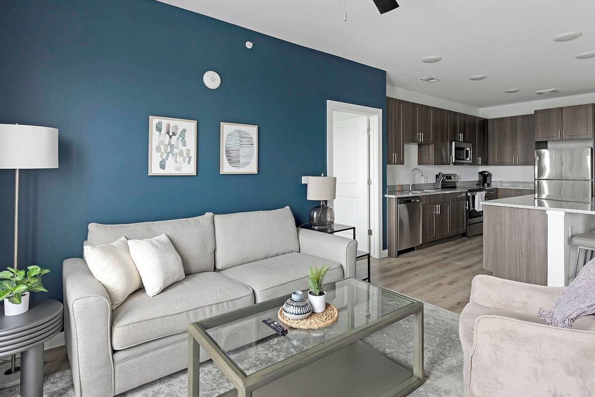 A modern living room with a grey sofa, a glass coffee table, and a kitchen in the background.