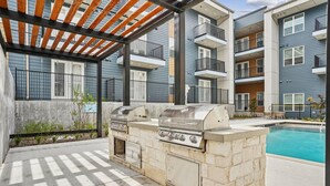 An outdoor barbecue area with a stone grill, a pergola, and a pool in the background. - Landing Apartments Garland, TX (Garland)