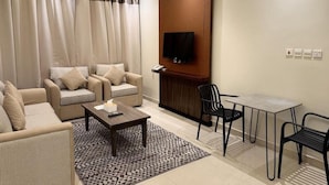 A hotel room with a sofa set, a coffee table, a TV mounted on a wooden panel, and a small dining table with chairs. - Nozol Aram 4 Elnafal (Riyadh)