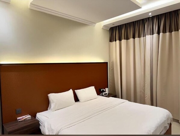 Una habitación de hotel con una cama grande, un cabecero de madera, una mesita de noche con lámpara y una ventana con cortinas.