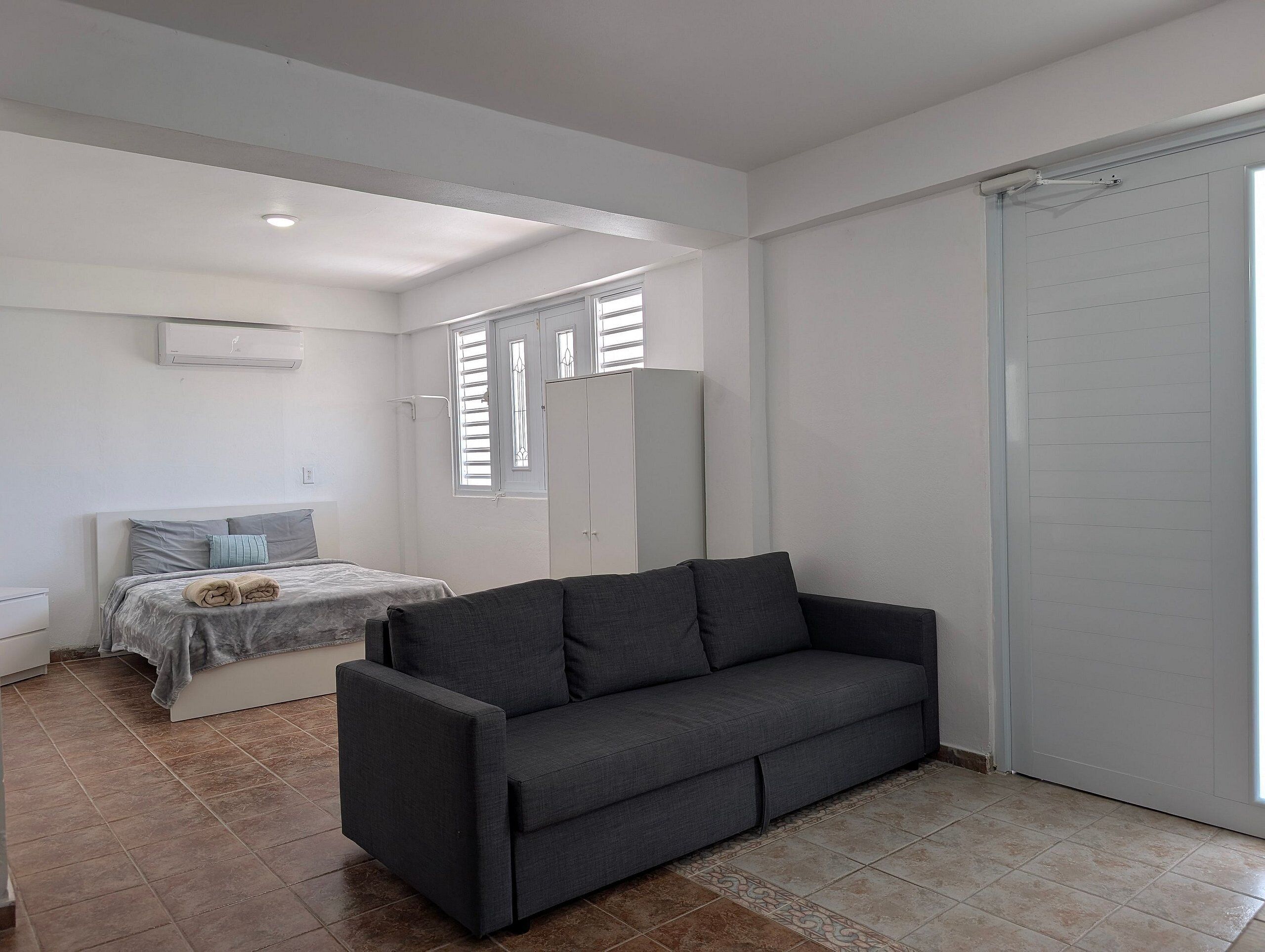A room with a bed, a dark sofa, a wardrobe, and a window with blinds.