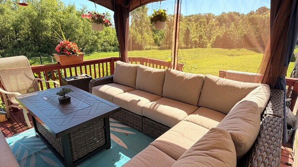 Une terrasse extérieure avec un canapé, une table, des meubles en osier, des plantes suspendues et une vue sur un paysage verdoyant.