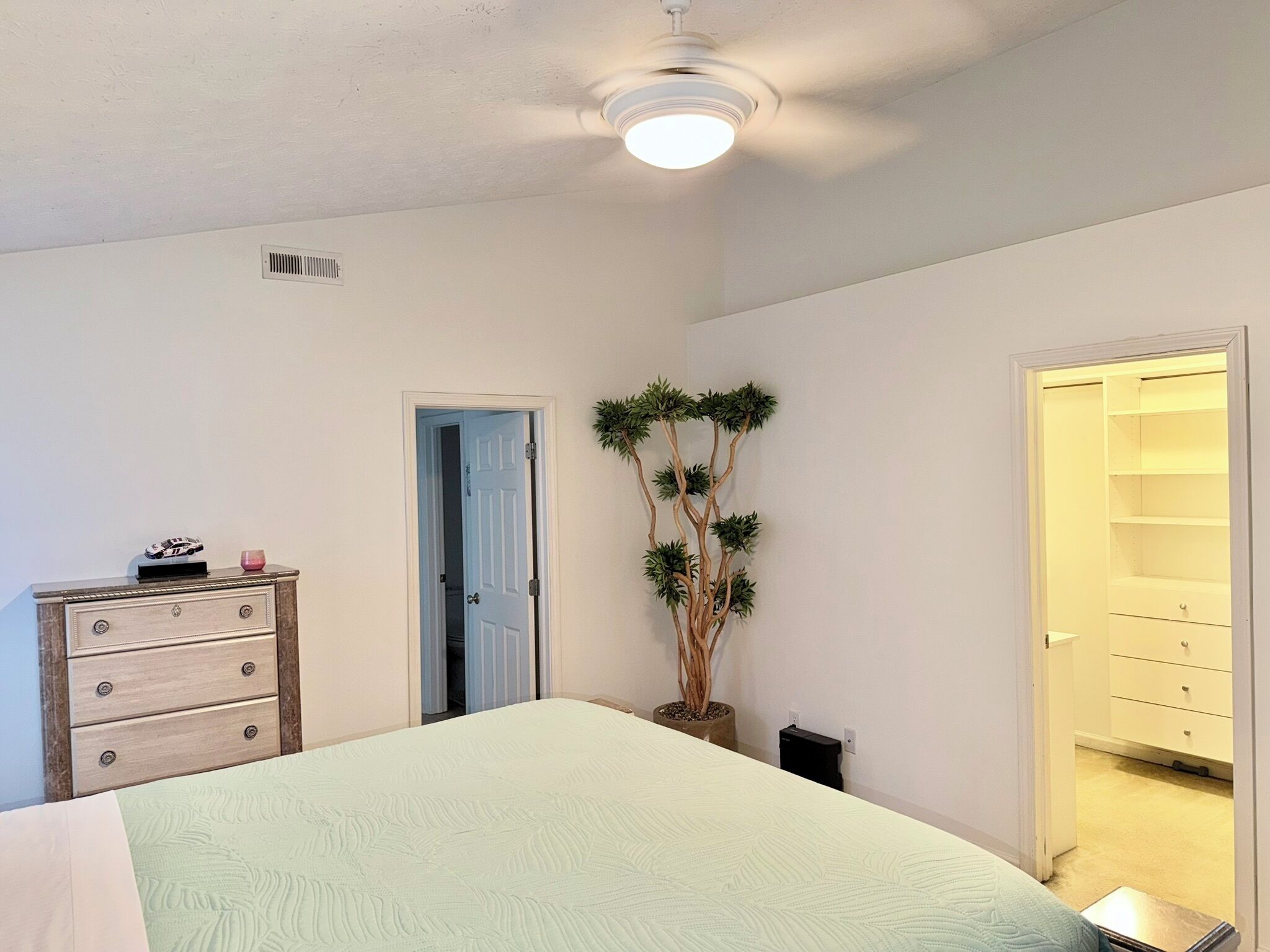 Une chambre à coucher comprenant un lit, une commode, une plante en pot, un placard et un ventilateur de plafond.