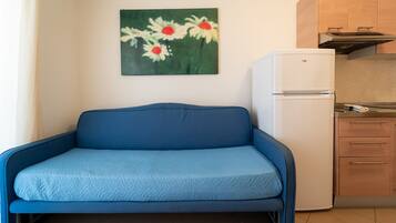 Une pièce de taille modeste, avec un canapé bleu, une petite kitchenette et un tableau représentant des fleurs accroché au mur.