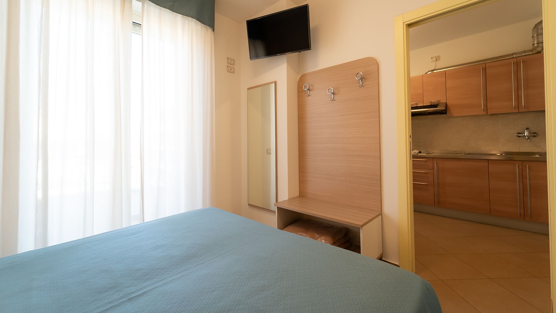 Une chambre à coucher comprenant un lit, un téléviseur fixé au mur, un miroir et une kitchenette équipée de placards et d’un évier.
