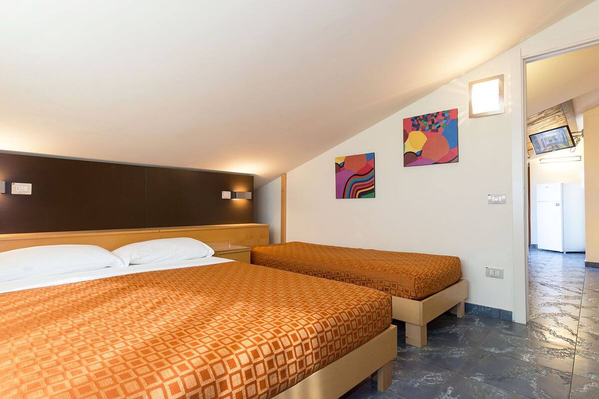 Hotellihuone, jossa on kaksi sänkyä, sängynpääty, seinään kiinnitetyt valaisimet ja taideteoksia.