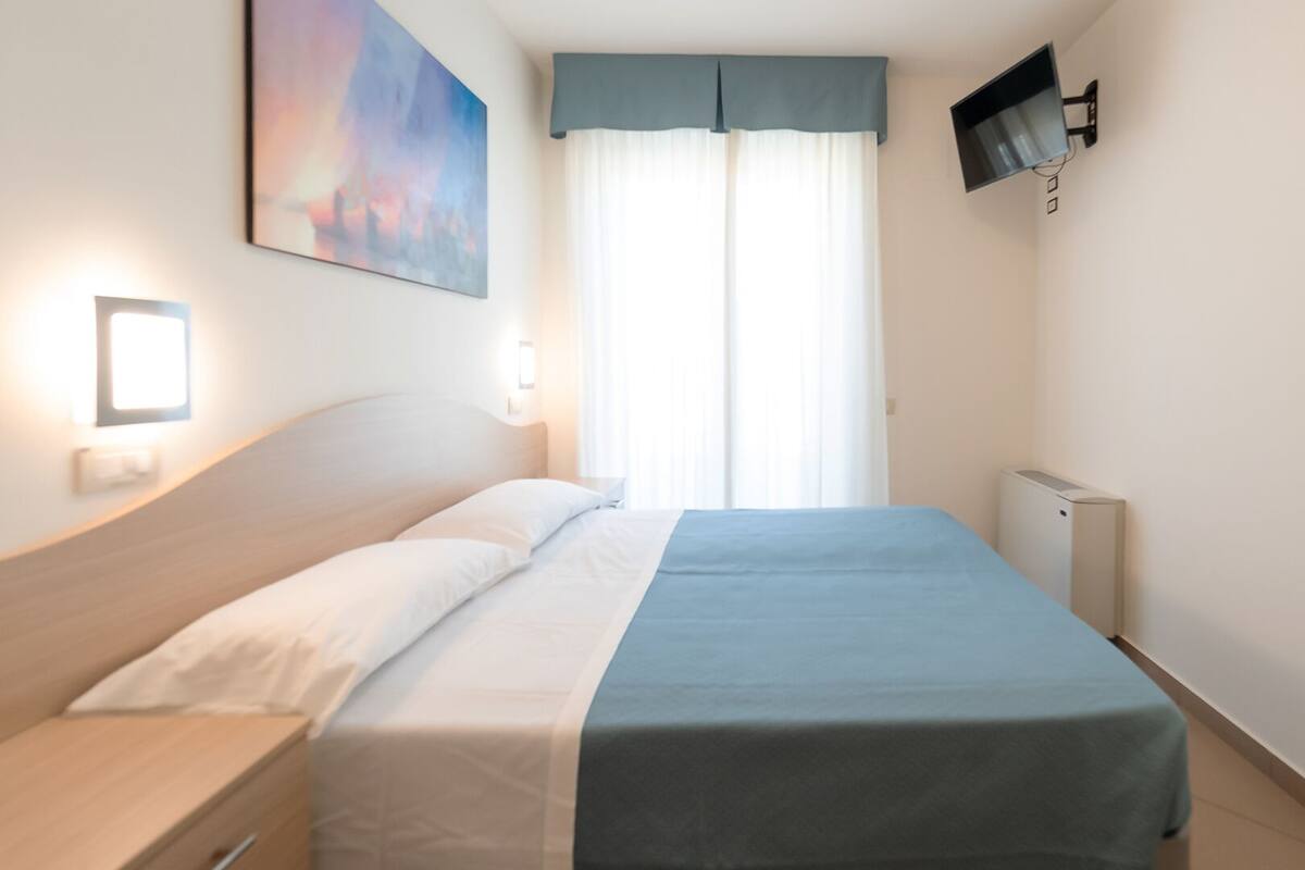 Hotellihuone, jossa on sänky, seinälle asennettu taulutelevisio, yöpöytä ja ikkuna, jossa on verhot.