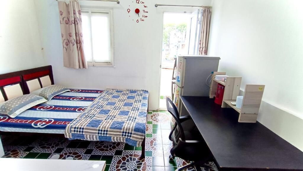 A small, clean room with a bed, a desk, and a chair. There is a window with curtains, a clock on the wall, and a small shelf with items.