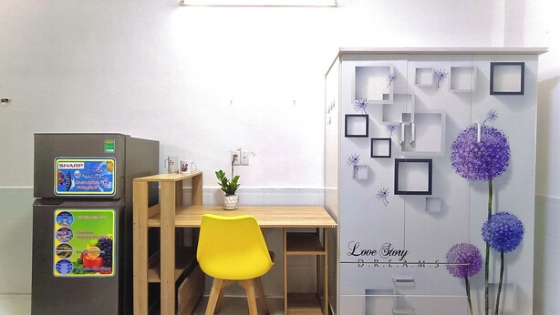 Ein Zimmer mit einem Holztisch, einem gelben Stuhl, einem Kühlschrank mit Sharp-Logo sowie einer Wand mit gerahmten Bildern und violetten Blumen.