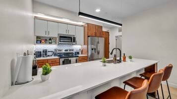 A modern kitchen with white cabinetry, a stainless steel refrigerator, a microwave, and a large island with bar stools.