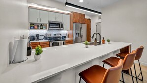 A modern kitchen with white countertops, stainless steel appliances, and wooden cabinets. There are three bar stools and a small potted plant on the counter.