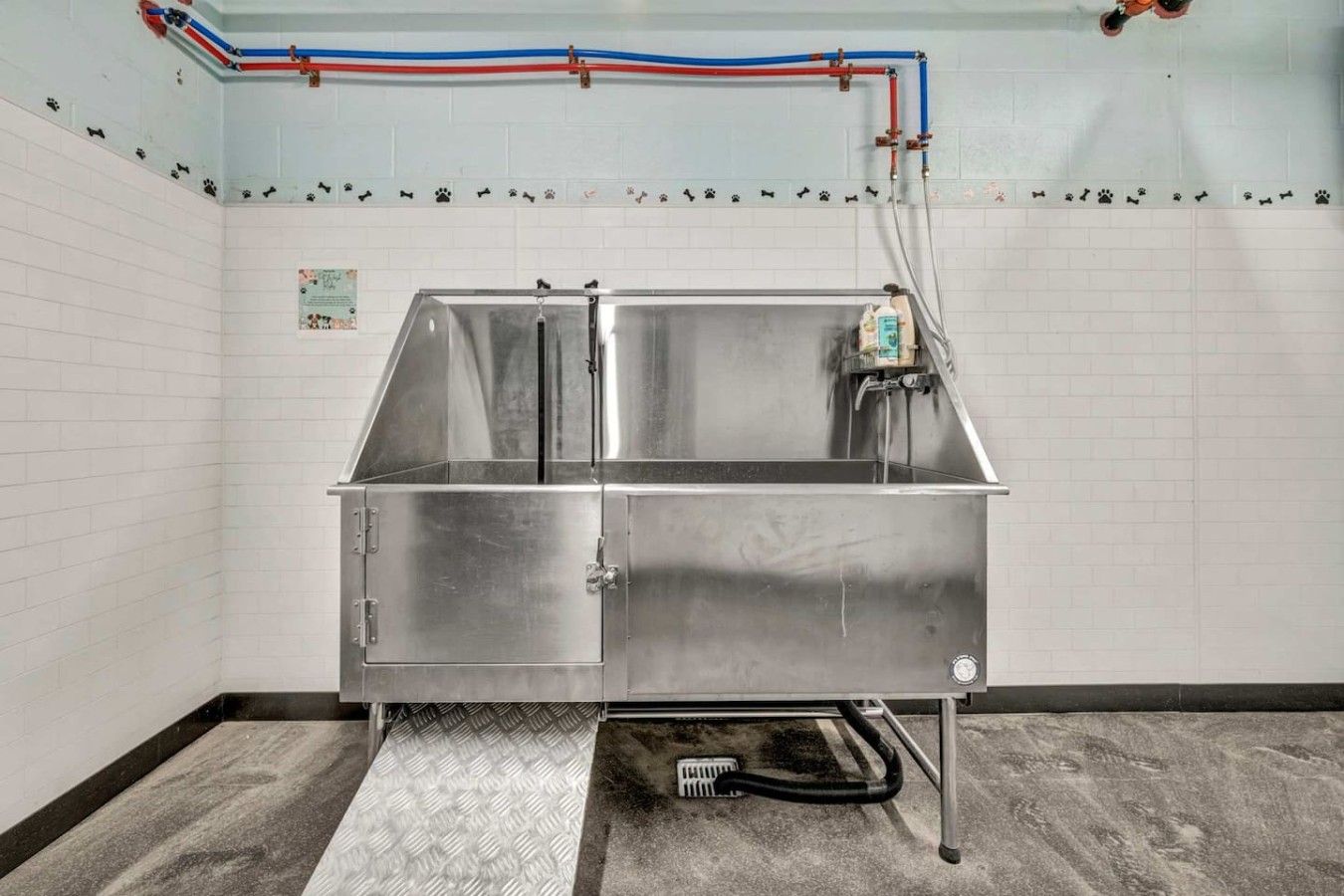 A stainless steel sink with a drainage hose, a wall-mounted faucet, and a tiled wall with a white baseboard.