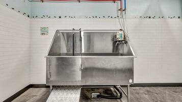 A stainless steel sink with a drainage hose, a wall-mounted faucet, and a tiled wall with a white baseboard.