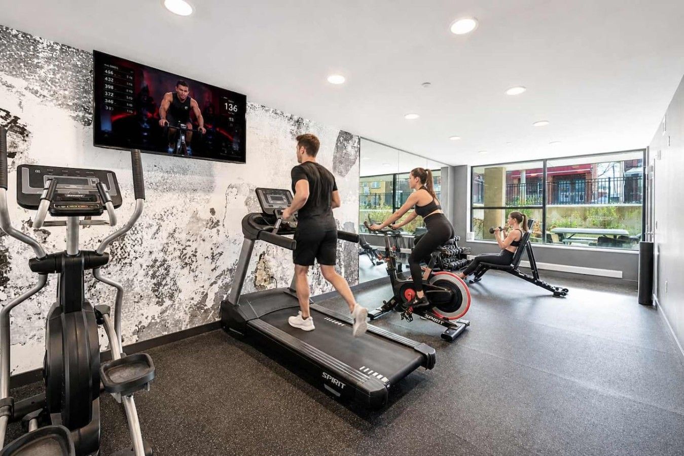 A gym with treadmills, an elliptical, and a stationary bike. There is a TV on the wall displaying a sports event.