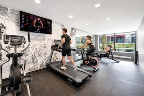 Une salle de sport équipée de tapis de course, d’un vélo elliptique et d’un vélo d’appartement. Un écran de télévision fixé au mur diffuse un événement sportif.