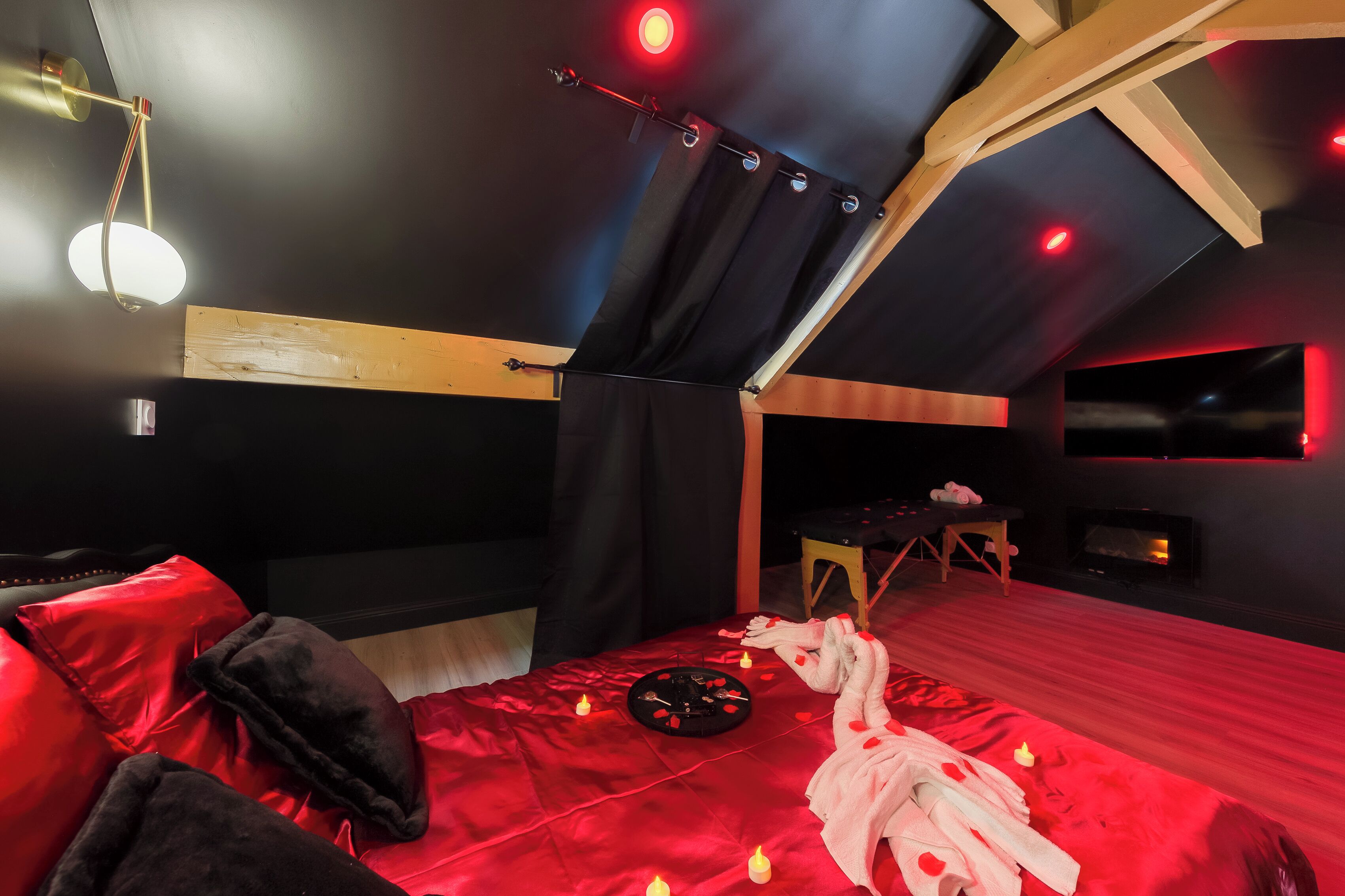 Ein schwach beleuchteter Raum mit einem Bett, roter Bettwäsche, Kerzen und einem kleinen Tisch mit einem Handtuch und einer Flasche.