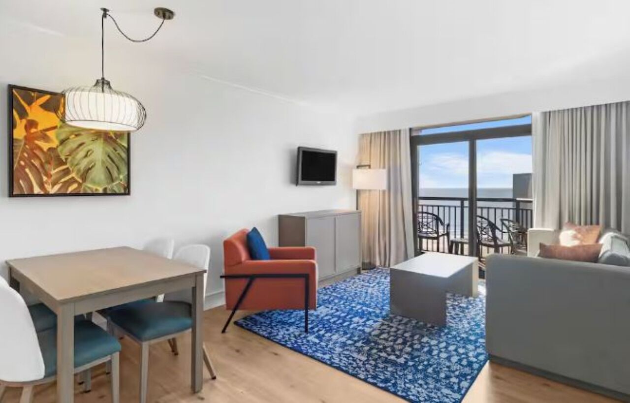 A modern hotel room with a dining table, a sofa, a TV, and a balcony with a view.
