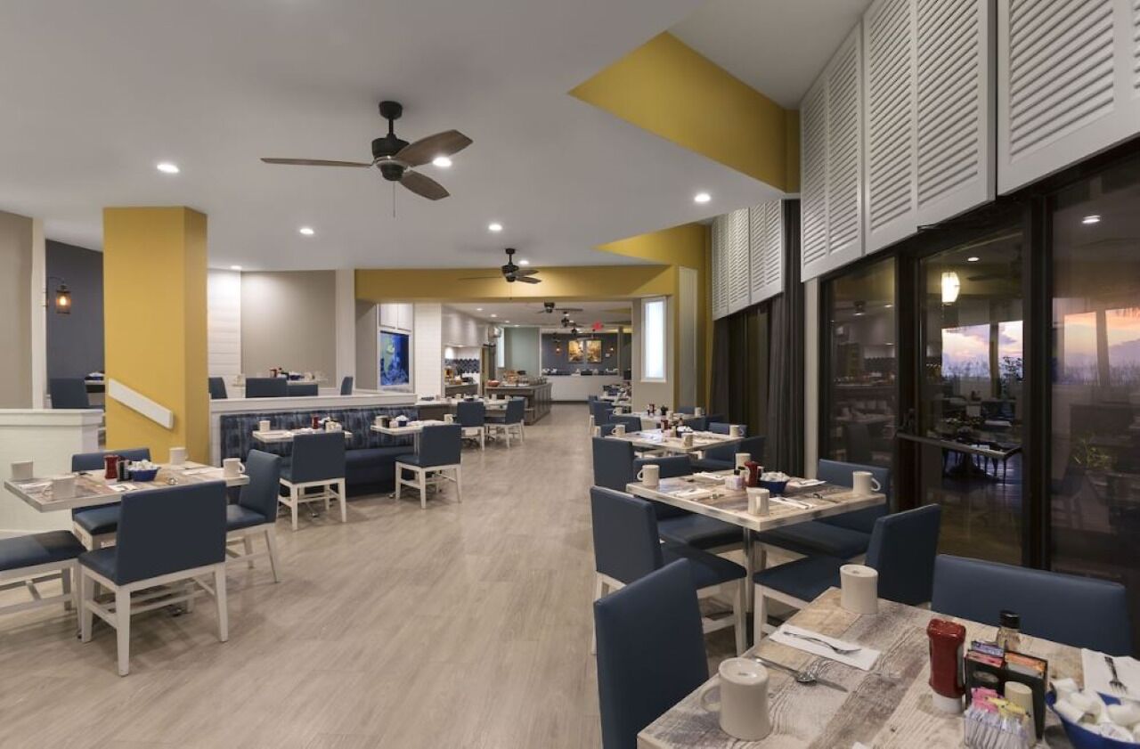 Un espace repas moderne, doté de plusieurs tables et chaises, de ventilateurs de plafond et offrant une vue sur l’extérieur grâce à de grandes fenêtres.