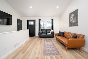 A modern living room with a wooden floor, a black leather sofa, an orange leather sofa, a black door, a TV mounted on the wall, and a framed artwork.