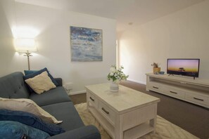 A living room with a sofa, a coffee table, a TV, a painting, and a lamp. - Stylish 2 BDR Apt Near QUT And RBWH With Parking (Kelvin Grove)
