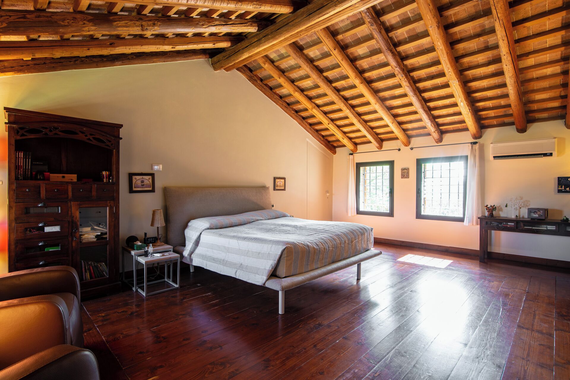 Un dormitorio con techo de madera, una cama, una mesita de noche, una estantería y dos ventanas.