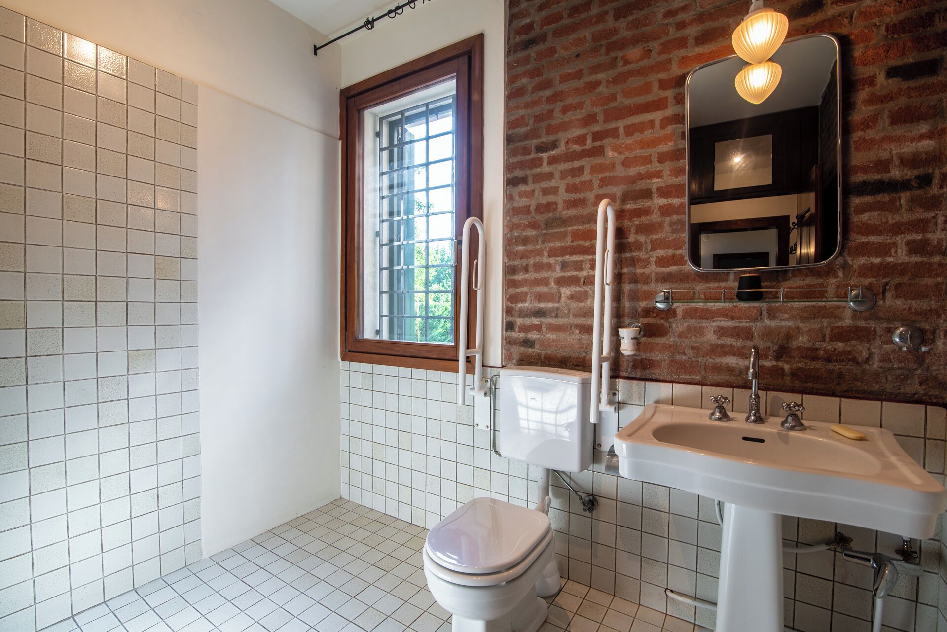 Un baño con inodoro blanco, un lavamanos con espejo arriba y una ventana con marco de madera.