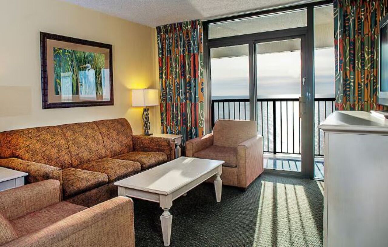 Une chambre d’hôtel avec un canapé, un fauteuil et une petite table. Elle dispose d’un balcon avec vue.