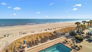 A beach view with a pool area, lounge chairs, and a clear sky.