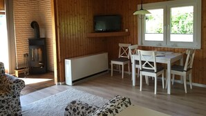 Een gezellig interieur van een blokhut met een open haard, een televisie bevestigd aan een houten balk, een eethoek met stoelen en een raam met uitzicht op groen.