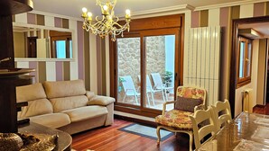 A living room with a beige sofa, a floral armchair, a dining table with chairs, a chandelier, and a view through large windows to the outside. - La Casa de Lorenzo, complete & practical accommodation. In Laguardia center! (Laguardia)