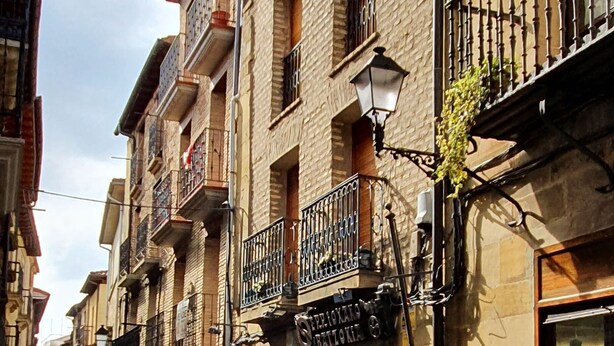 A narrow street lined with multi-story buildings featuring balconies, a shop on the ground floor, and a potted plant near the entrance. - La Casa de Lorenzo, complete & practical accommodation. In Laguardia center! (Laguardia)