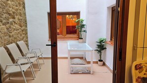 A narrow hallway with white walls, a stone wall on one side, and a wooden door at the end. There are several white chairs lined up against the wall and a small white table with a potted plant beside it. - La Casa de Lorenzo, complete & practical accommodation. In Laguardia center! (Laguardia)