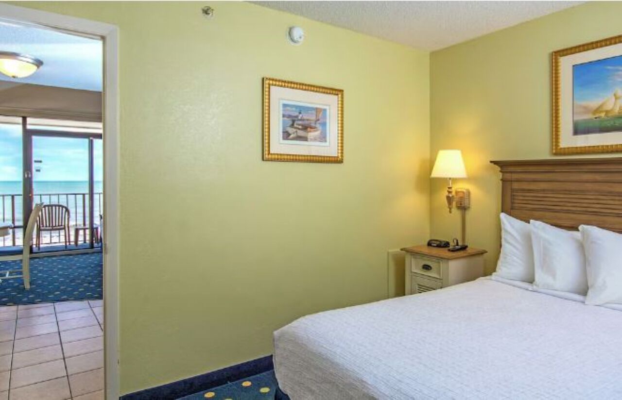 A hotel room with a bed, a nightstand, a lamp, a framed picture on the wall, and a view of the balcony.