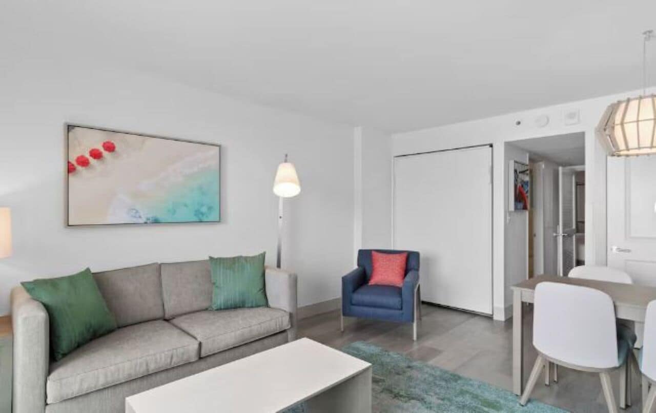 A modern living room with a sofa, a dining table, and a chair. There is a painting on the wall and a lamp.