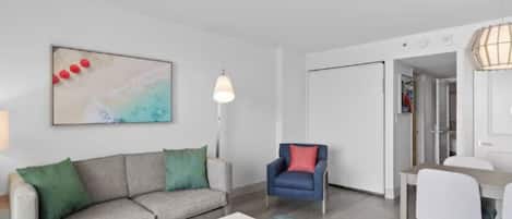 A modern living room with a sofa, a dining table, and a chair. There is a painting on the wall and a lamp.