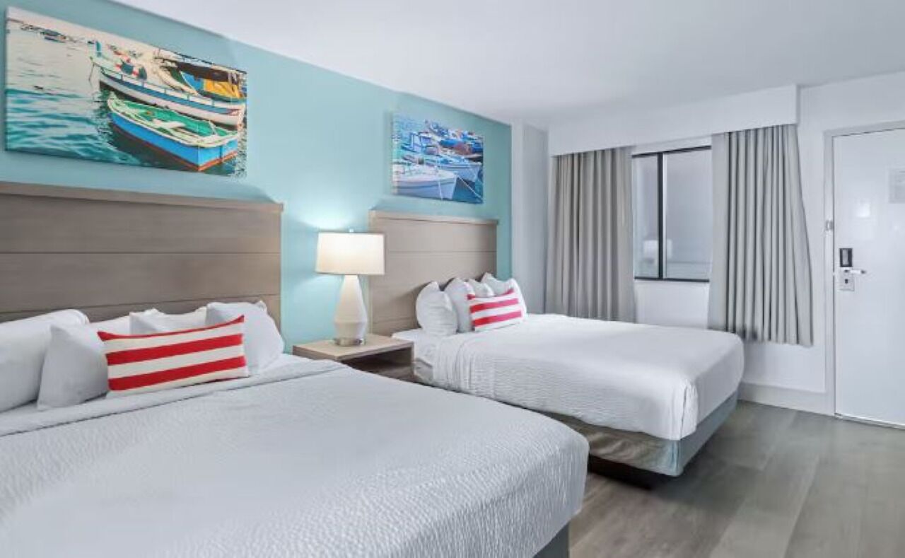 Une chambre d’hôtel avec deux lits, une table de chevet avec une lampe, une fenêtre avec des rideaux et des œuvres d’art accrochées aux murs.