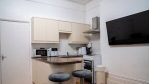 Eine kompakte Küche mit weißen Schränken, einer Mikrowelle, einer Kaffeemaschine und einem kleinen Backofen. Zwei Hocker und ein an der Wand befestigtes Fernsehgerät sind vorhanden.