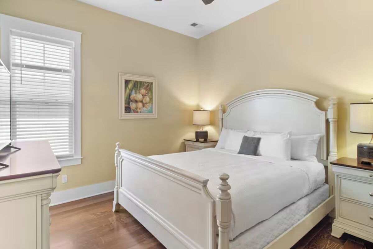 A bedroom with a large bed, a nightstand, a desk, and a window with blinds.