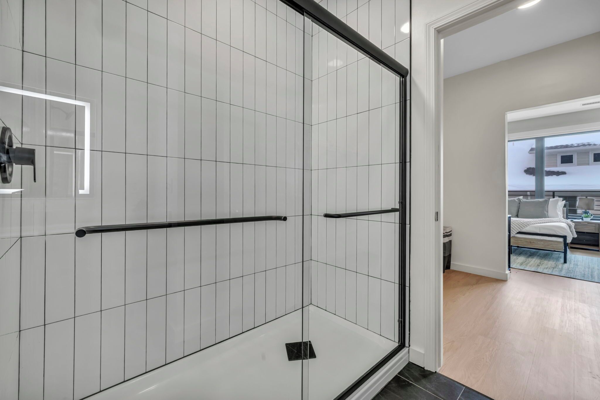 A modern bathroom with white tiled walls, a glass shower enclosure, and a visible bedroom with a bed and a nightstand.